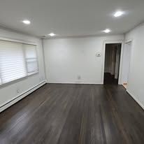 Empty living room with hardwood floors and white walls. Window with blinds on one side.