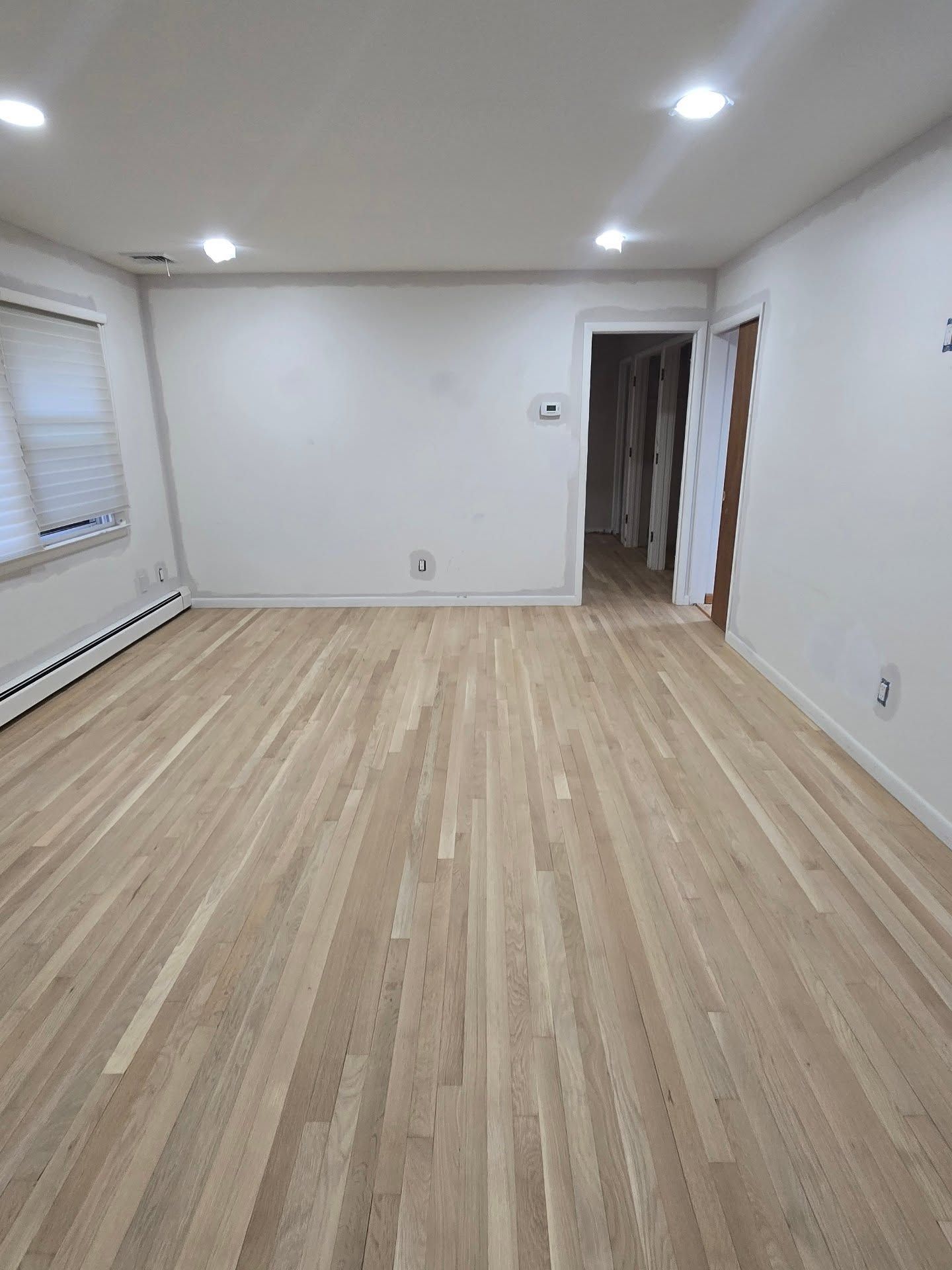 Empty room with light wood floors, white walls, and recessed lighting. A door leads to a hallway.