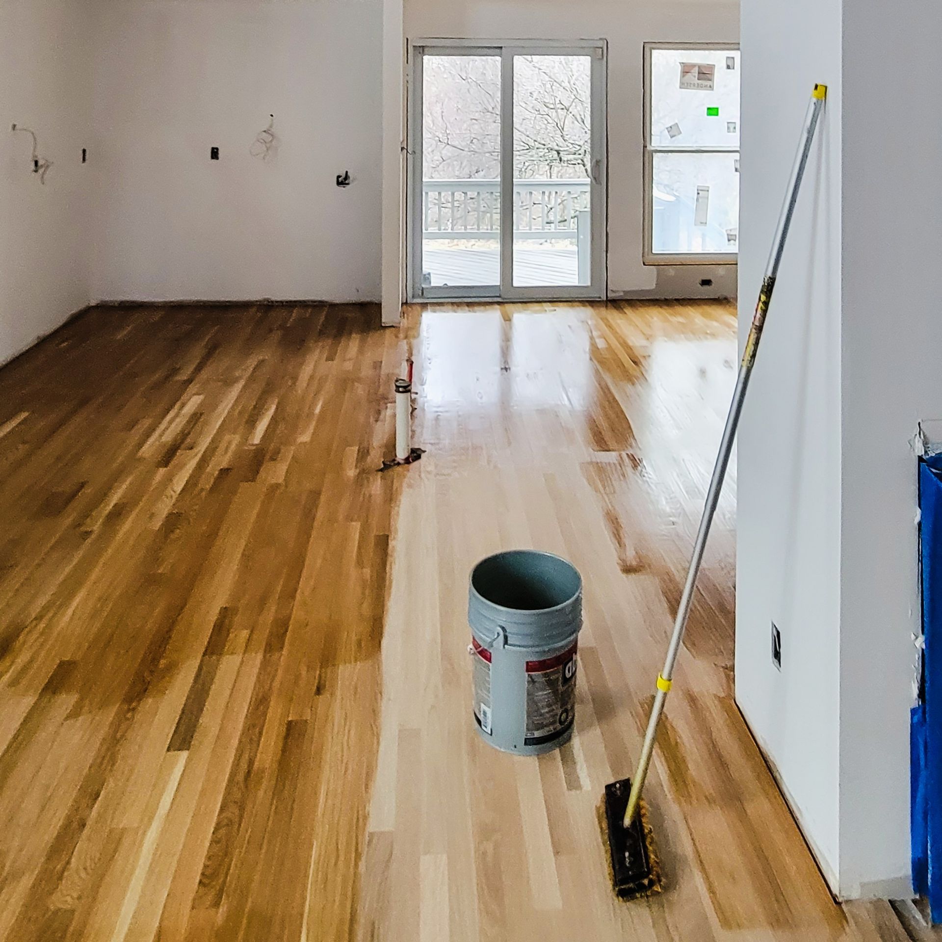 Wood flooring being stained; bucket and applicator visible in unfinished room.