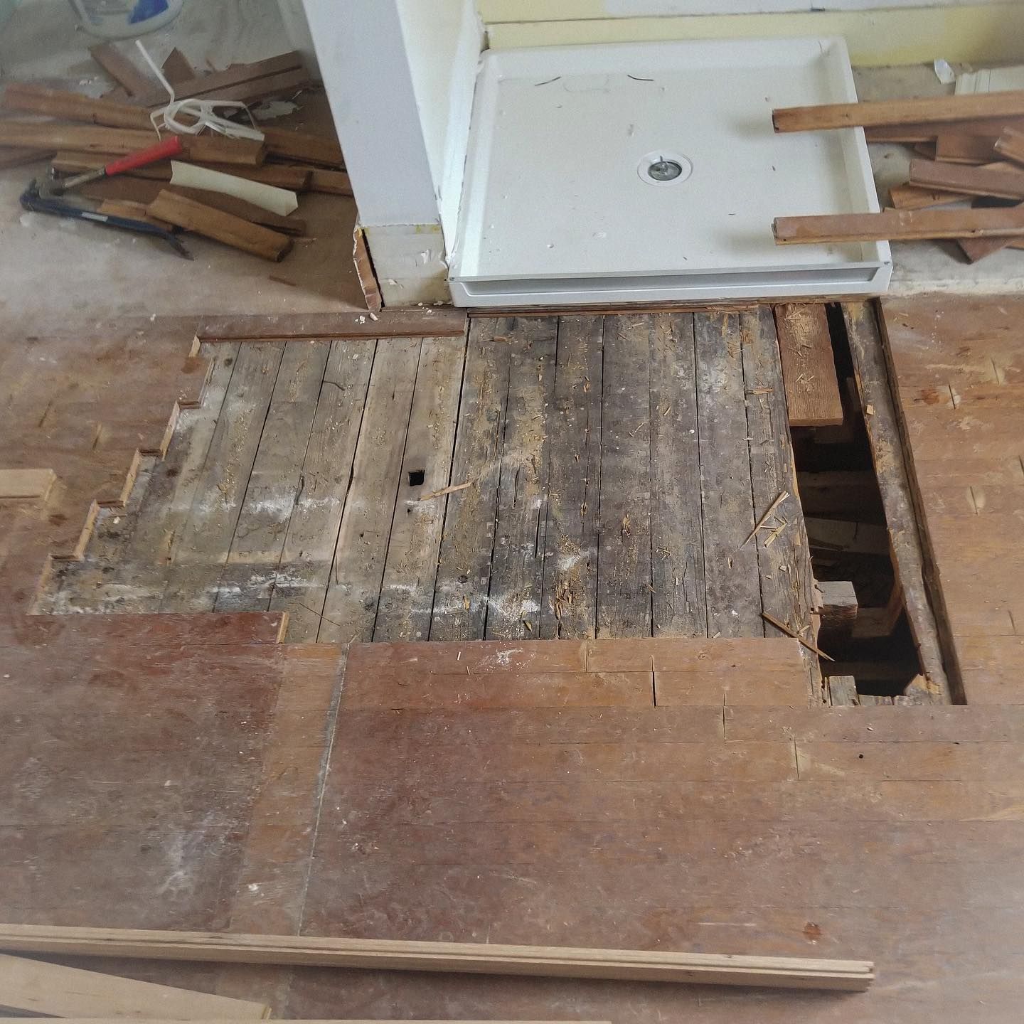Floor demolition in a bathroom; exposed subfloor next to a shower base and scattered wooden planks.