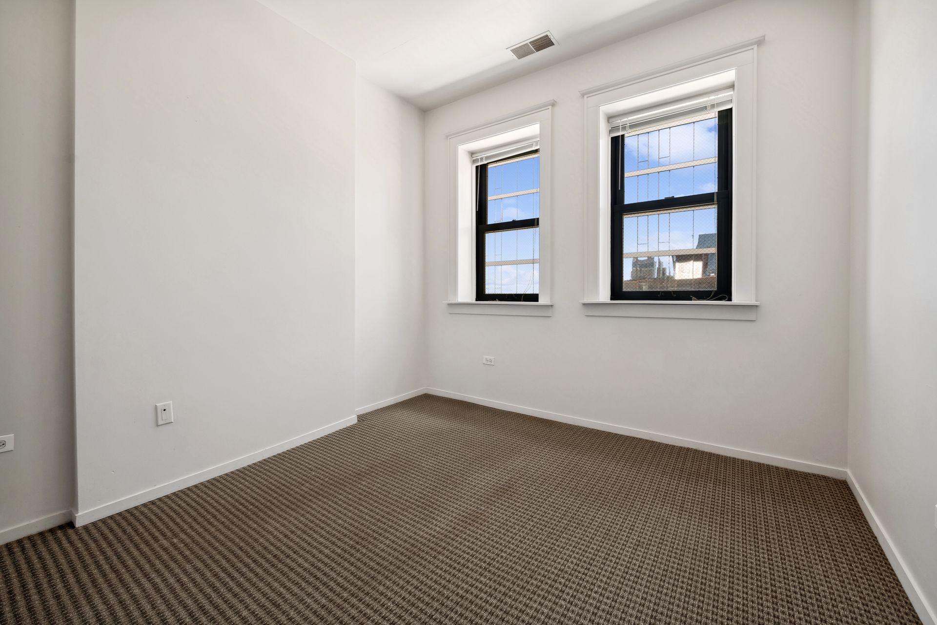 An empty room with two windows and a brown carpet.
