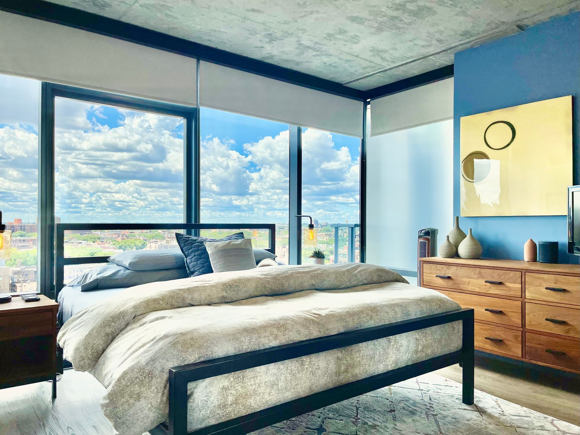 A bedroom with a bed , dresser , nightstand , and large windows.