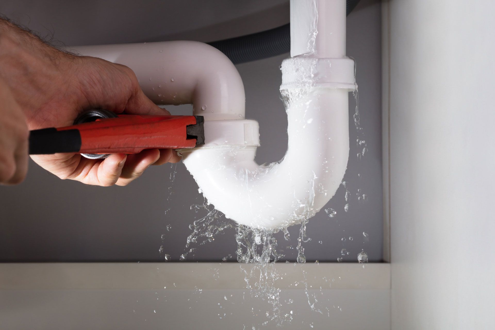 Hand With Wrench Attempts to Fix a Leaky White Pipe Under a Sink; Water Sprays — Full On Plumbing & Gas in Winnellie, NT