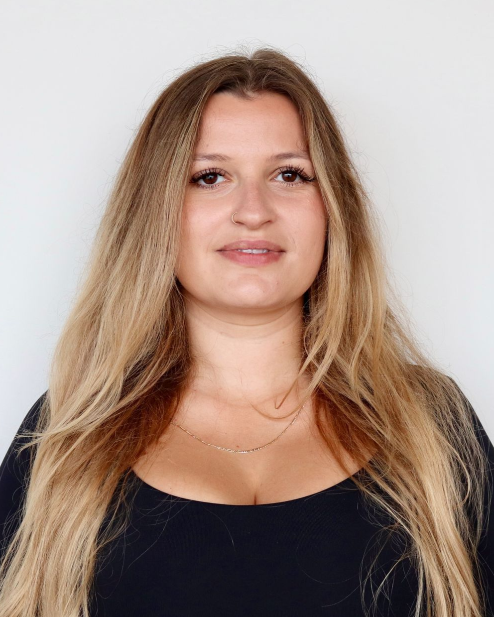 Woman with long, wavy blonde hair smiles, wearing a black top against a white wall.