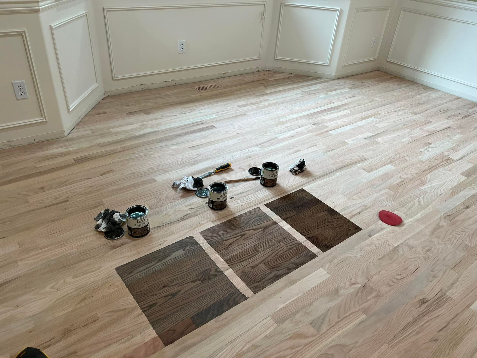 Wooden floor with stain samples. Paint cans and tools on floor. Light walls.