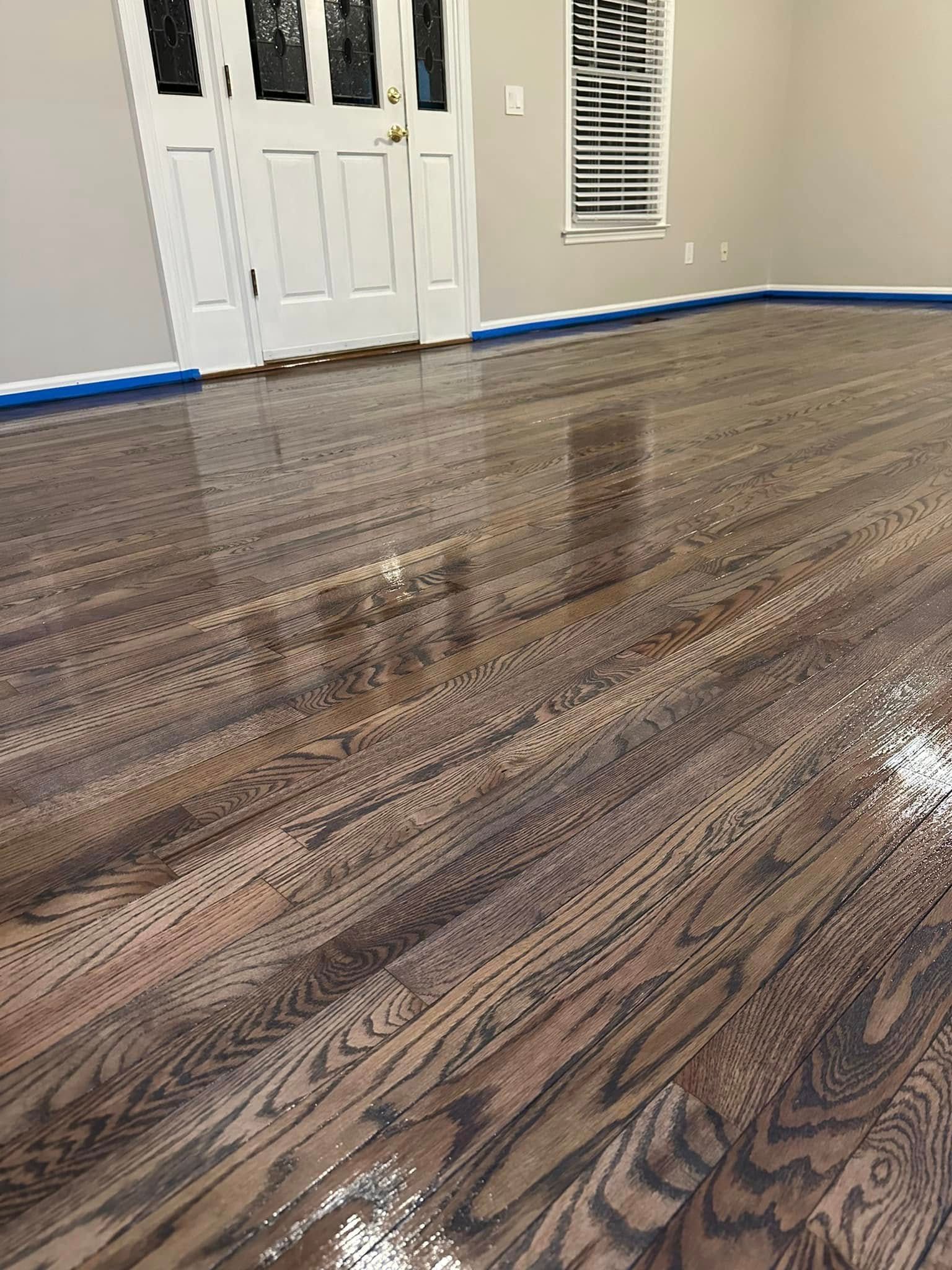 Glossy, newly-finished hardwood floor reflecting light. Doors and a window are in the background, edges taped off with blue tape.