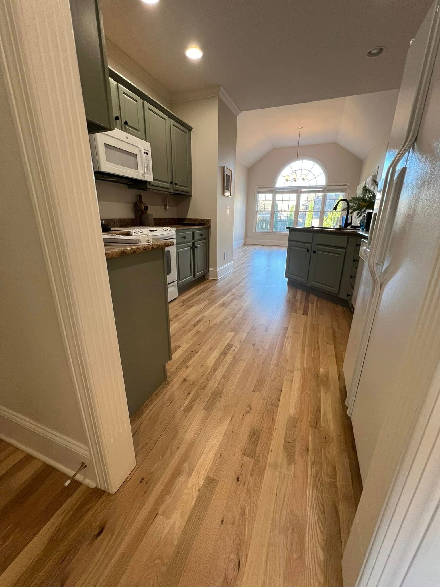 Kitchen with green cabinets, wood floors, and view to a light-filled living area.