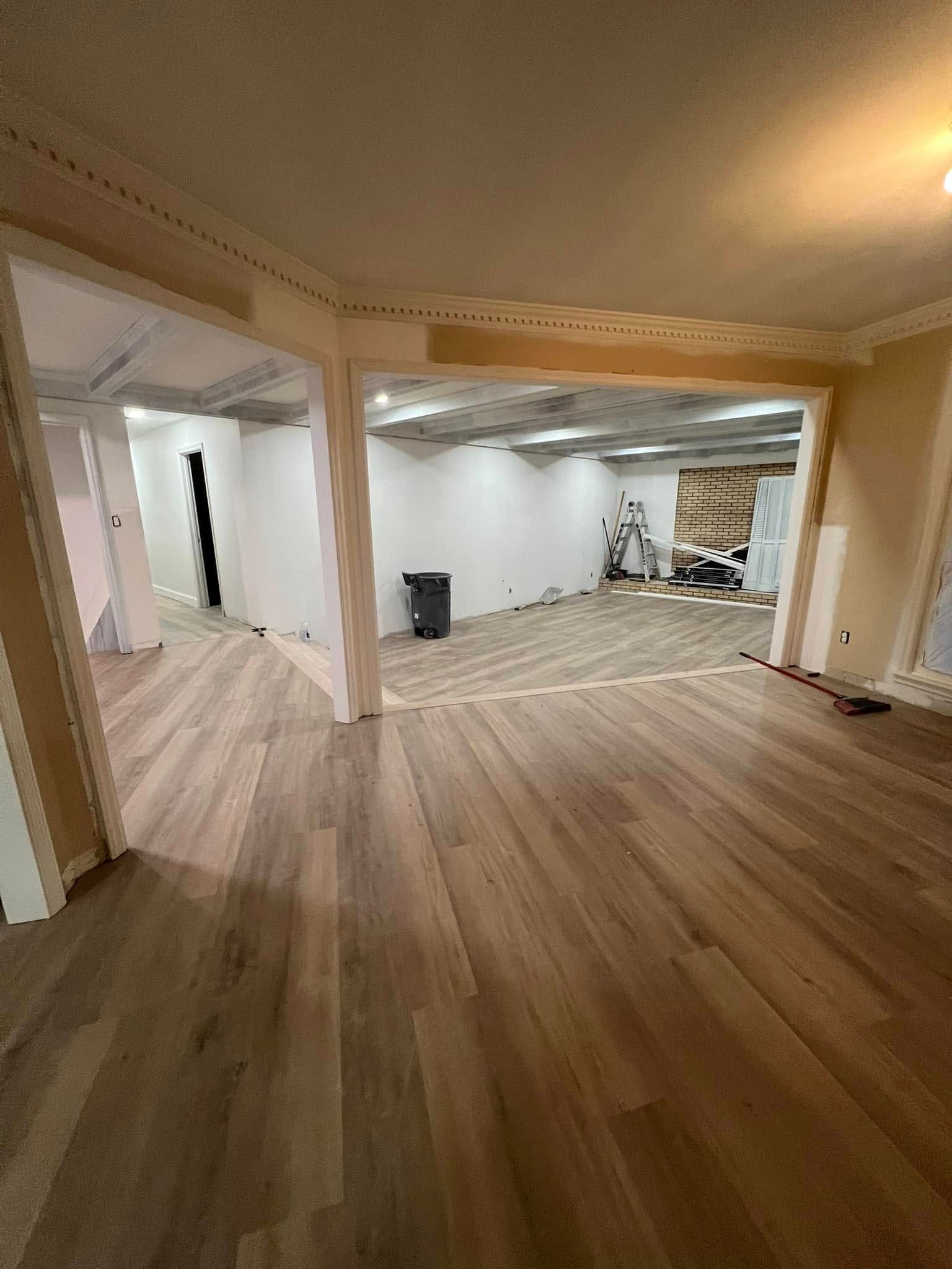 Interior view of a room under renovation with hardwood floors, white walls, and a large opening to another space.