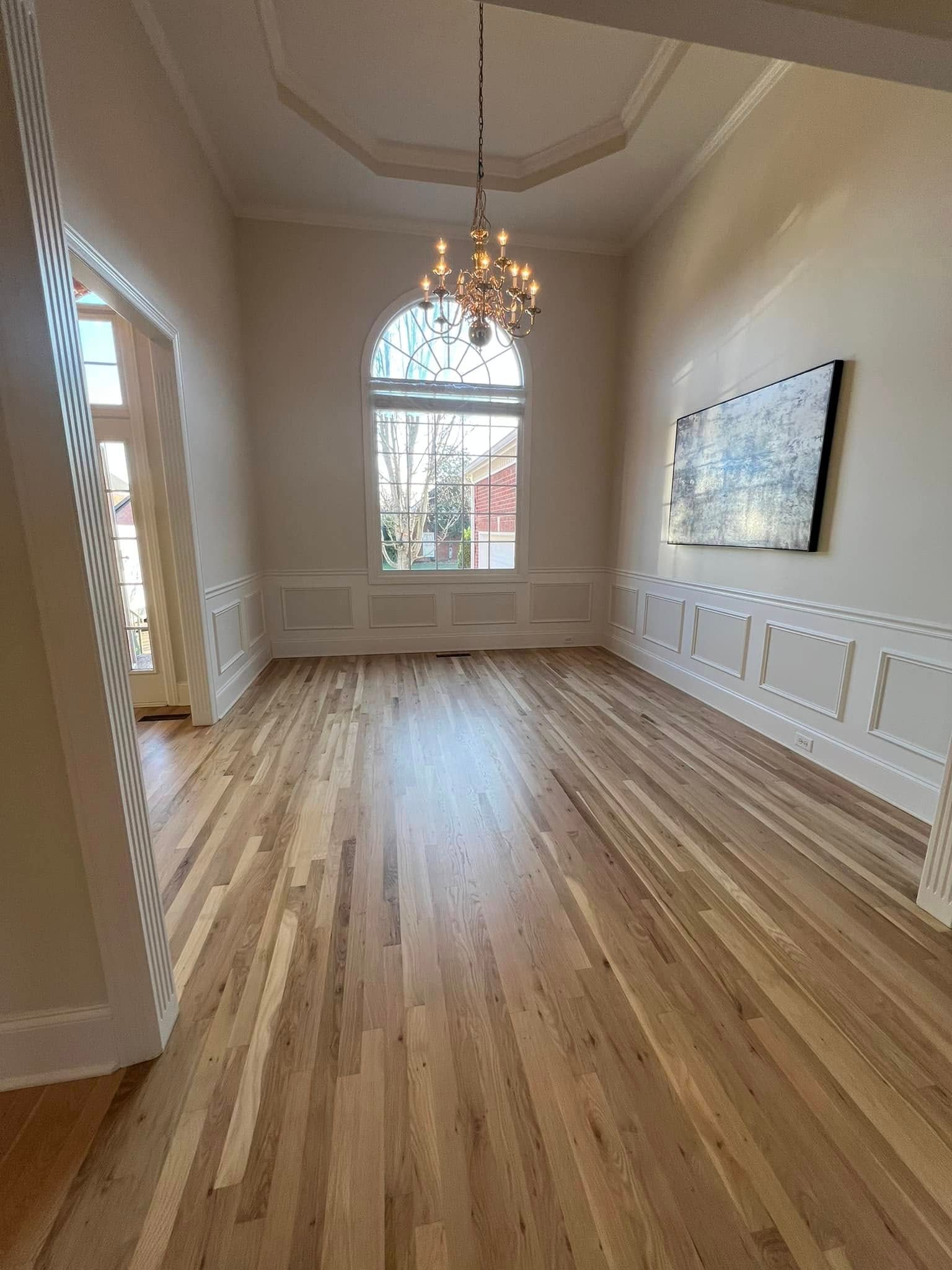 Empty room with hardwood floors, a large window, chandelier, and a framed artwork.