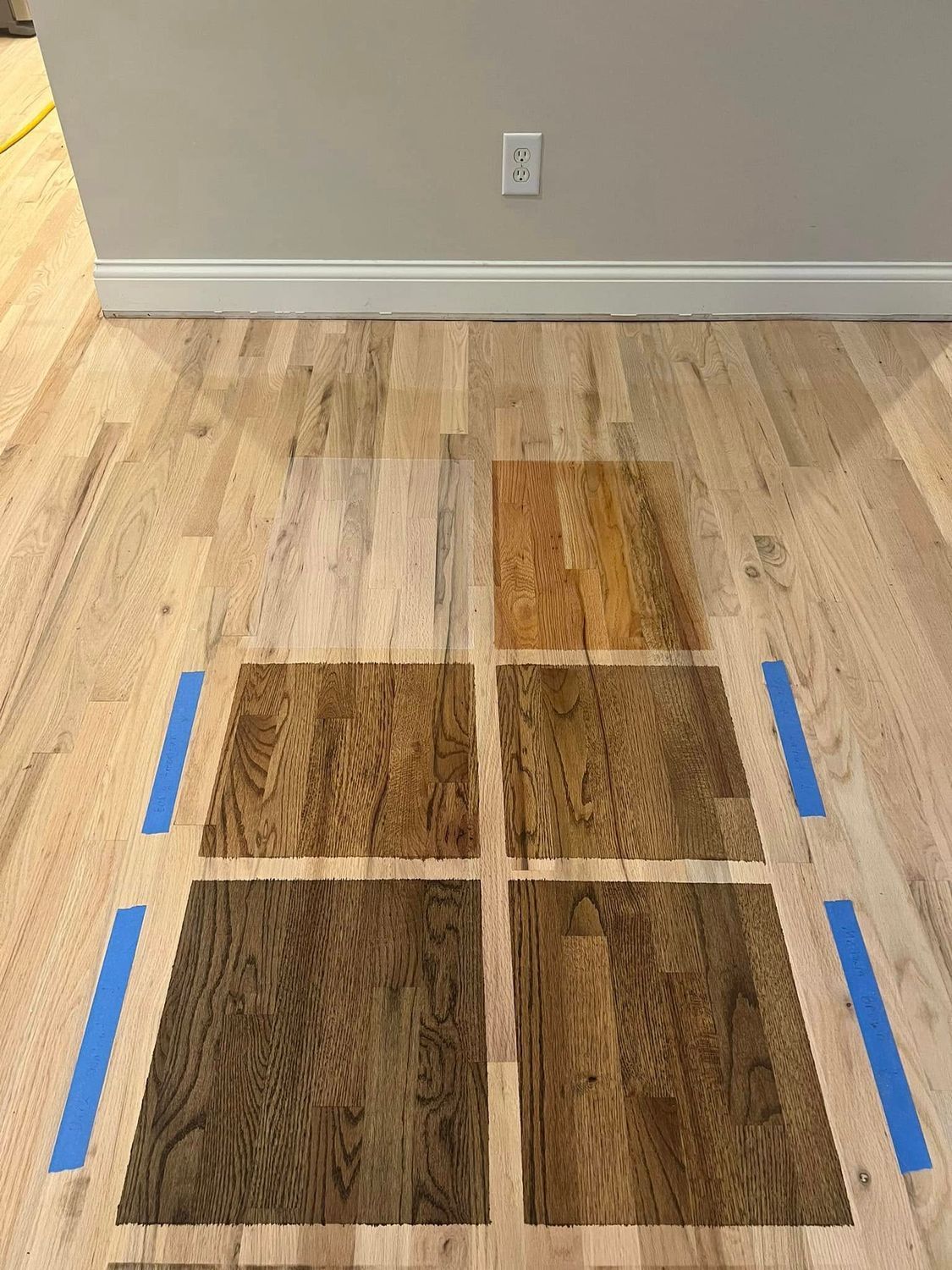 Hardwood floor with six stained samples in various shades of brown. Blue tape marks the boundaries.