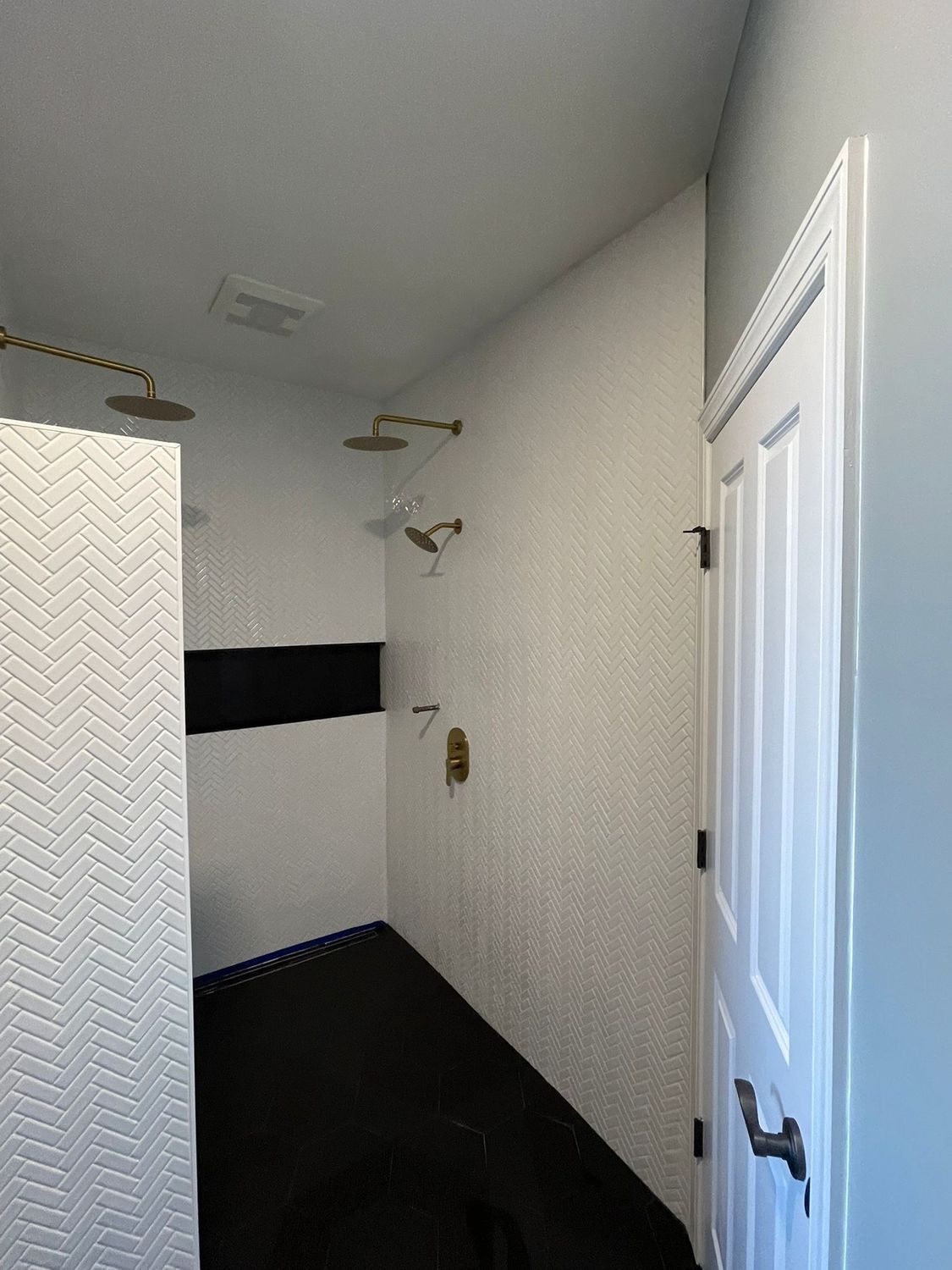 Modern shower with white tiled walls and black floor, gold shower fixtures, and a closed white door.