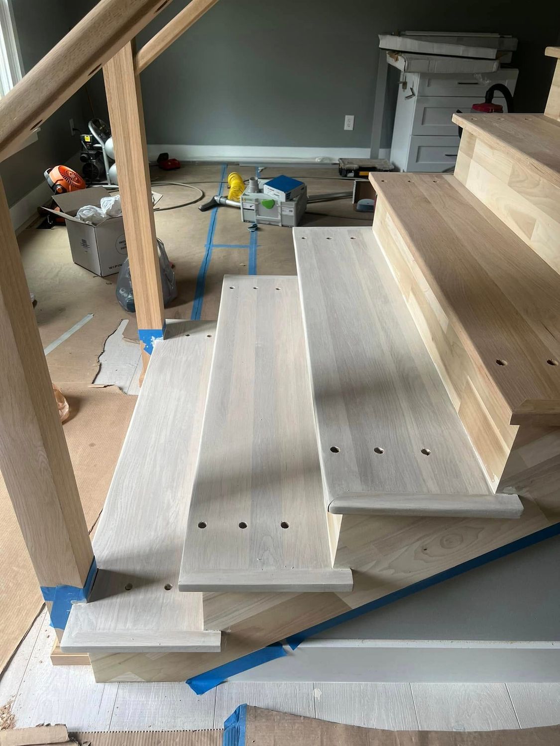 Wooden staircase under construction, with light-colored steps, posts, and railing. Blue tape marks the edges.
