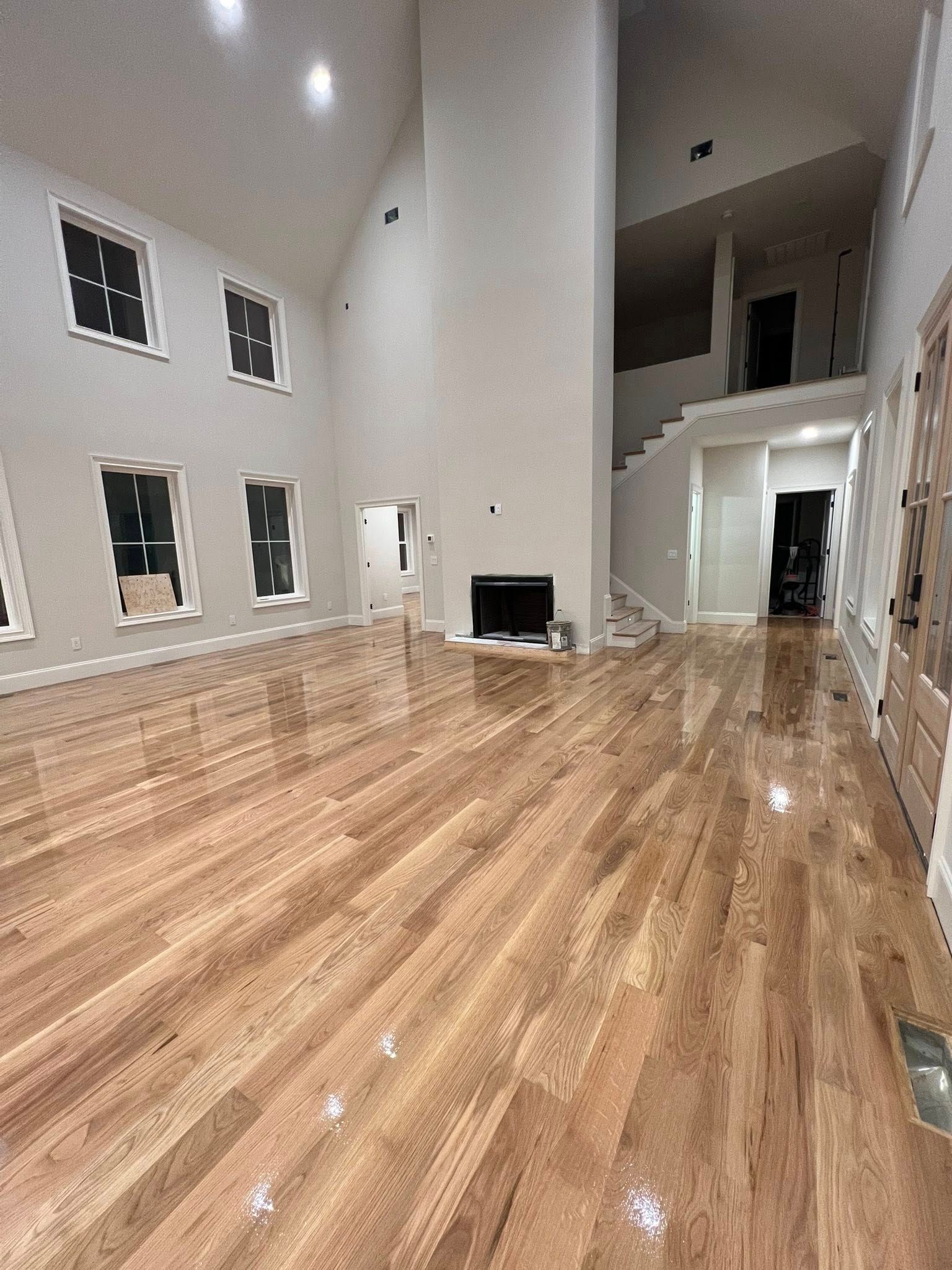 Spacious room with hardwood floors, fireplace, and staircase. High ceilings, several windows, and light-colored walls.