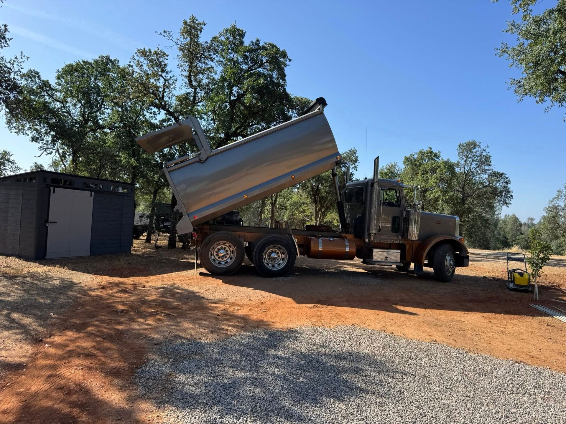Brown dump truck with raised bed, dumping material outdoors near a small structure. Sunny day.