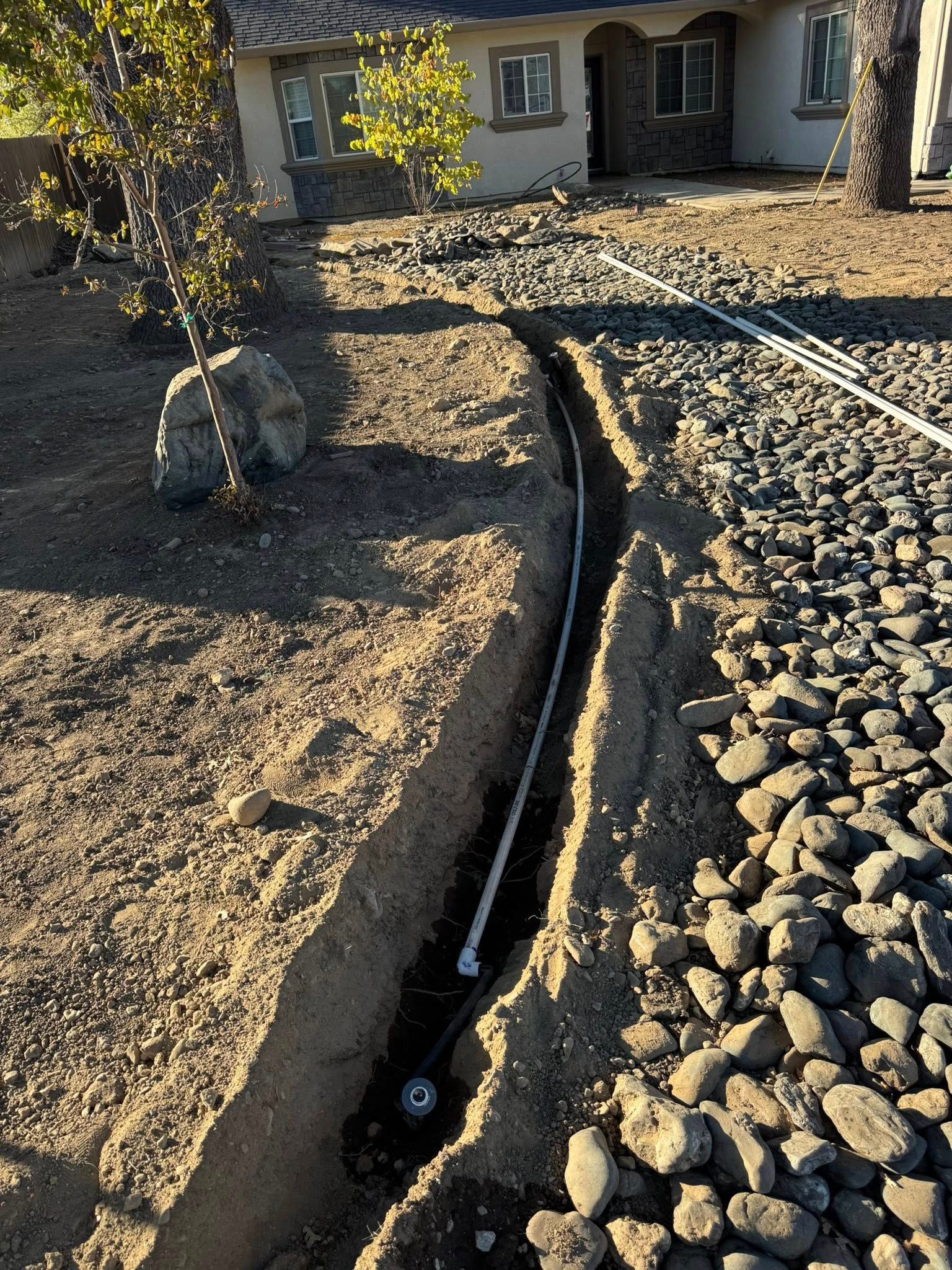 A trench dug in dirt, with white pipes laid inside; a house in the background and rocks on the right.