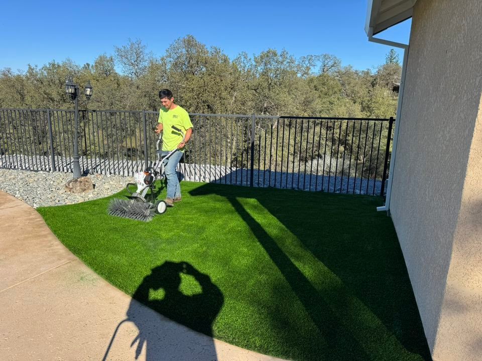 Man using a lawn sweeper on artificial turf, near a black fence, gravel, and a building on a sunny day.