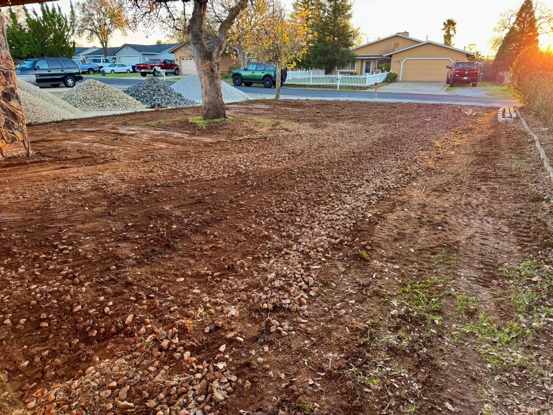 Cleared dirt yard with a pile of rocks in front of houses with cars parked on the street.