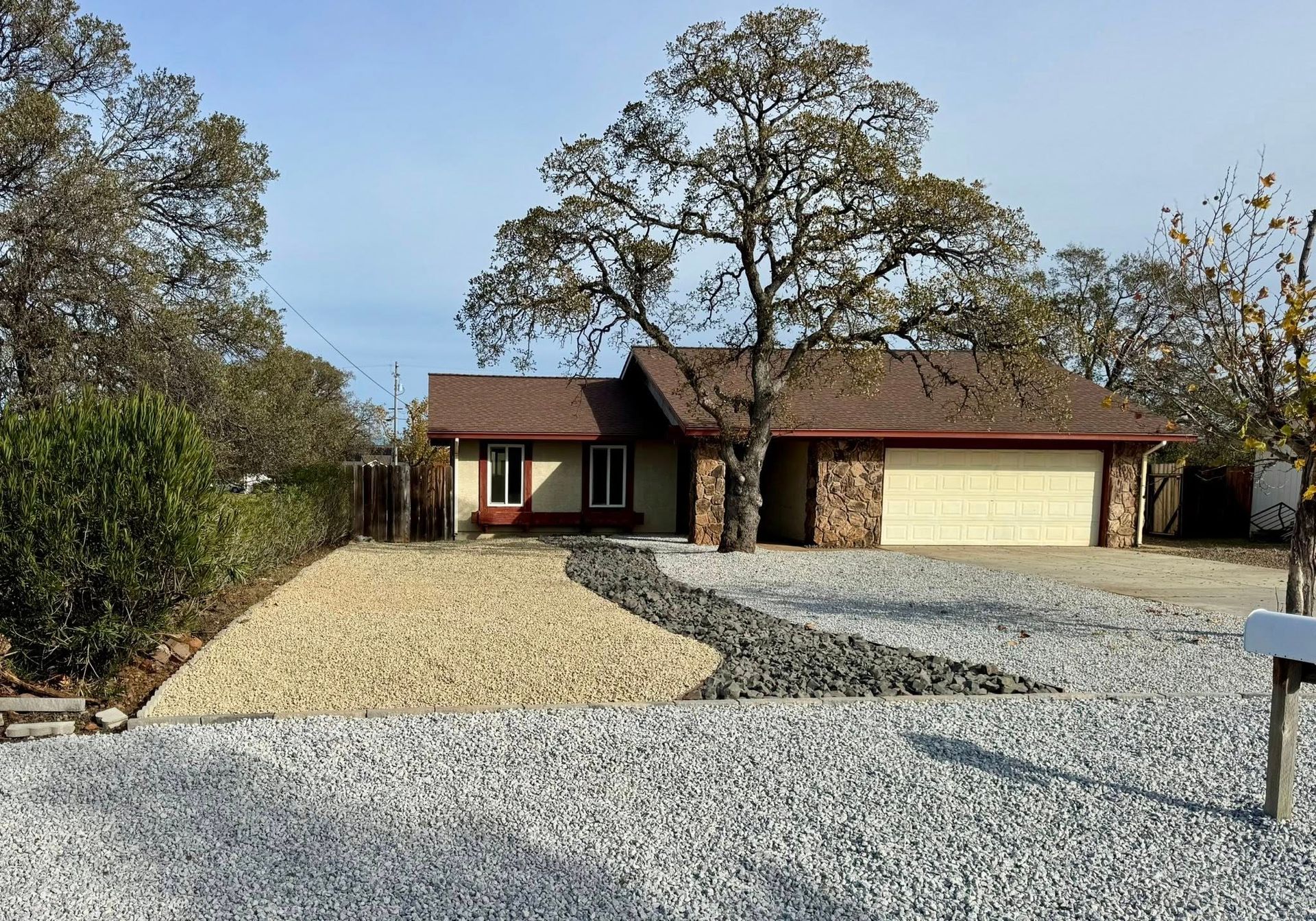 Tan house with a stone facade and attached garage, nestled among trees. Gravel landscaping leads to the front door.