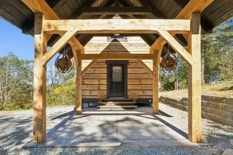 Wooden timber frame porch covering a cabin's entrance; hanging decorative globes, gravel driveway, stone wall.
