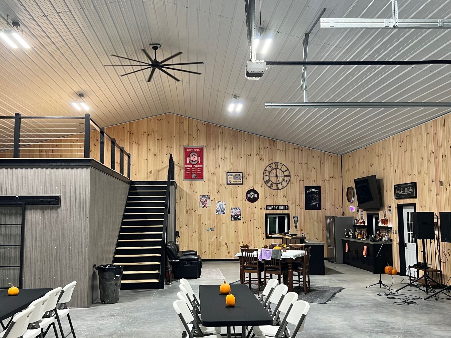 Interior of a large venue with wooden walls, a black staircase, tables, chairs, and a loft area.