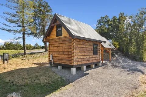 Wooden cabin with a corrugated metal roof, on concrete blocks, next to a gravel driveway.