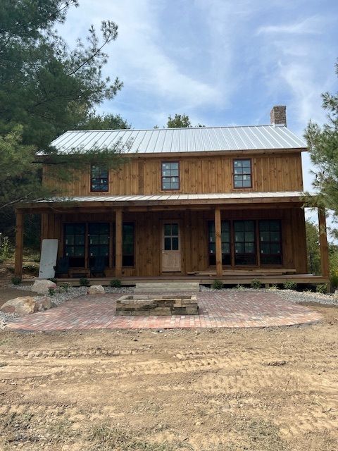 Two-story wooden house with a porch and a metal roof, situated on a brick patio with trees in the background.