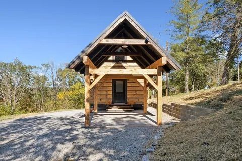 Rustic cabin with wooden beams under a clear blue sky. Gravel driveway and grassy hillside.