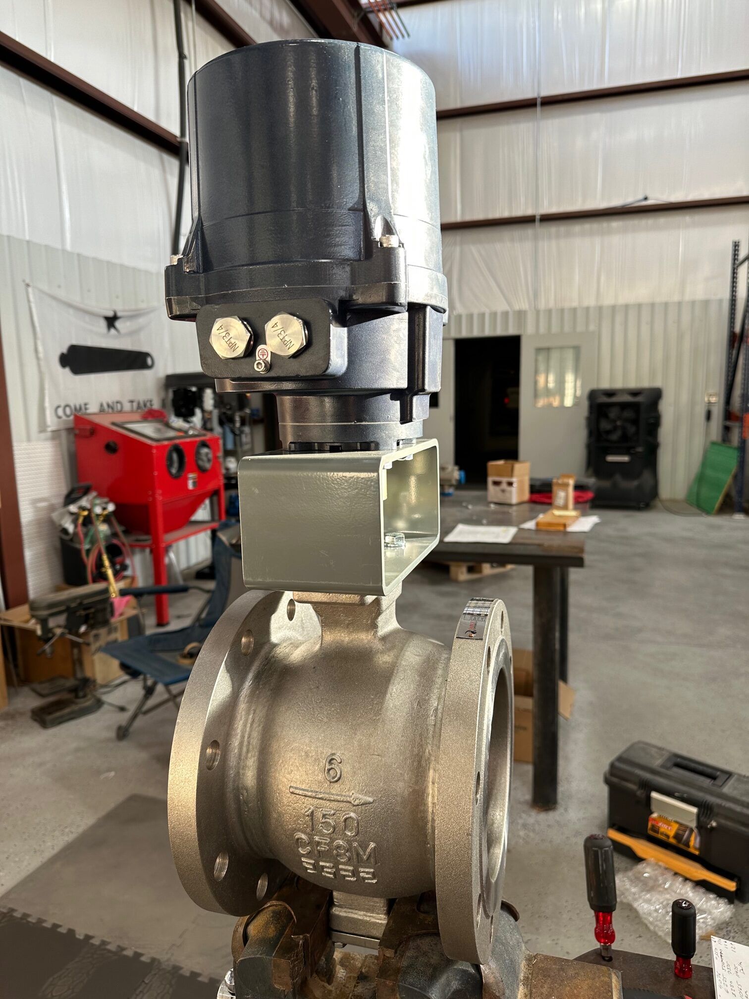 An industrial metal ball valve with a dark gray electric actuator mounted on top, sitting on a workbench in a shop.