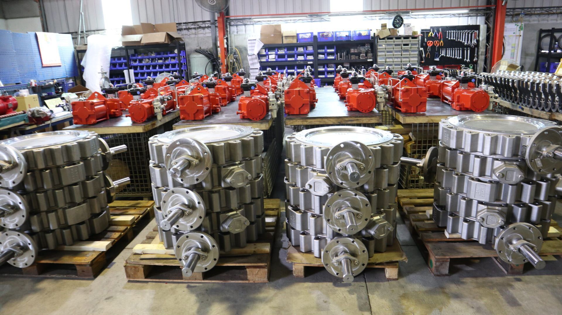 Stacks of metallic industrial valves on wooden pallets in a warehouse, with rows of orange components in the background.