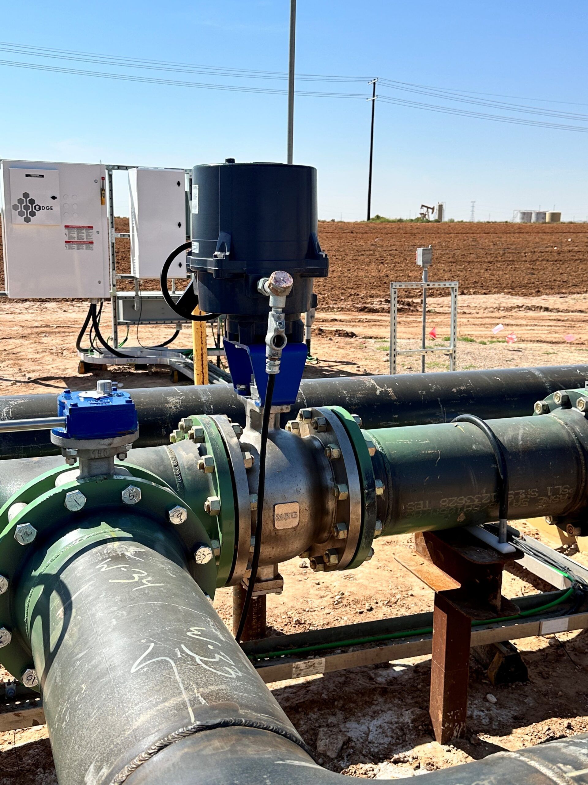 A large black motorized valve attached to industrial piping in a sandy, outdoor oil field setting under a clear blue sky.