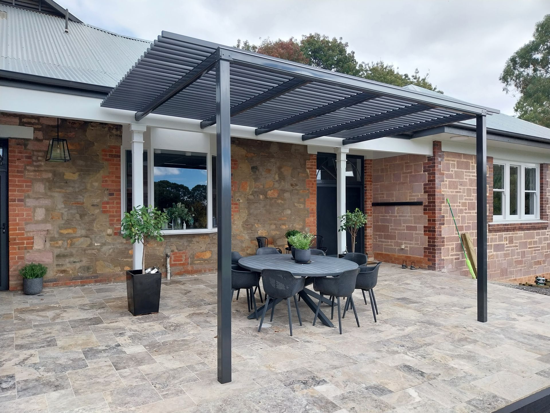 White farmhouse porch with wooden deck, chairs, and red flower pots, surrounded by trees.