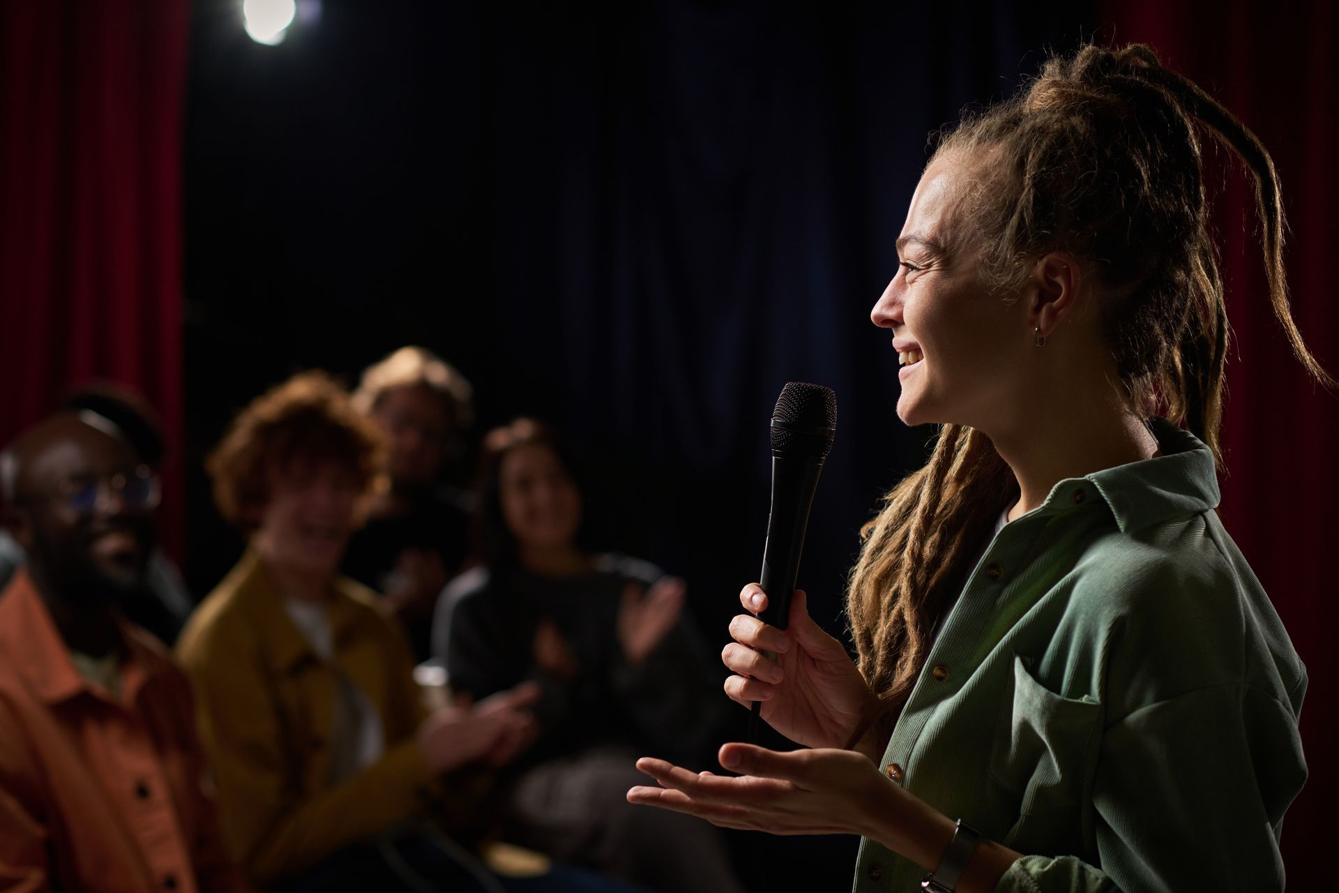 Woman with dreadlocks speaks into a microphone, addressing a seated audience in a theater setting.