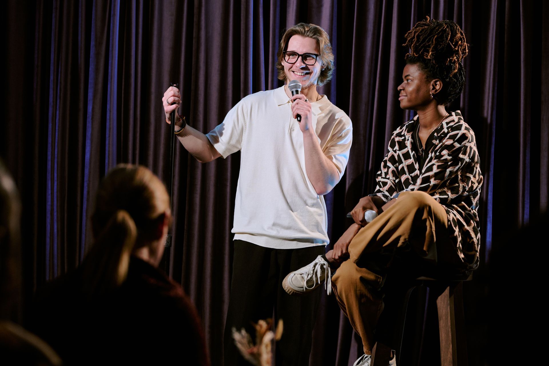 Man with microphone speaks. Seated woman listens on stage. Dim setting, audience in foreground.