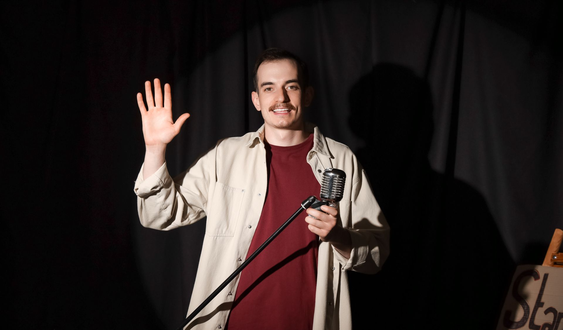 Man on a stage waves with one hand, holding a microphone. He wears a beige shirt over a maroon shirt, with a black backdrop.