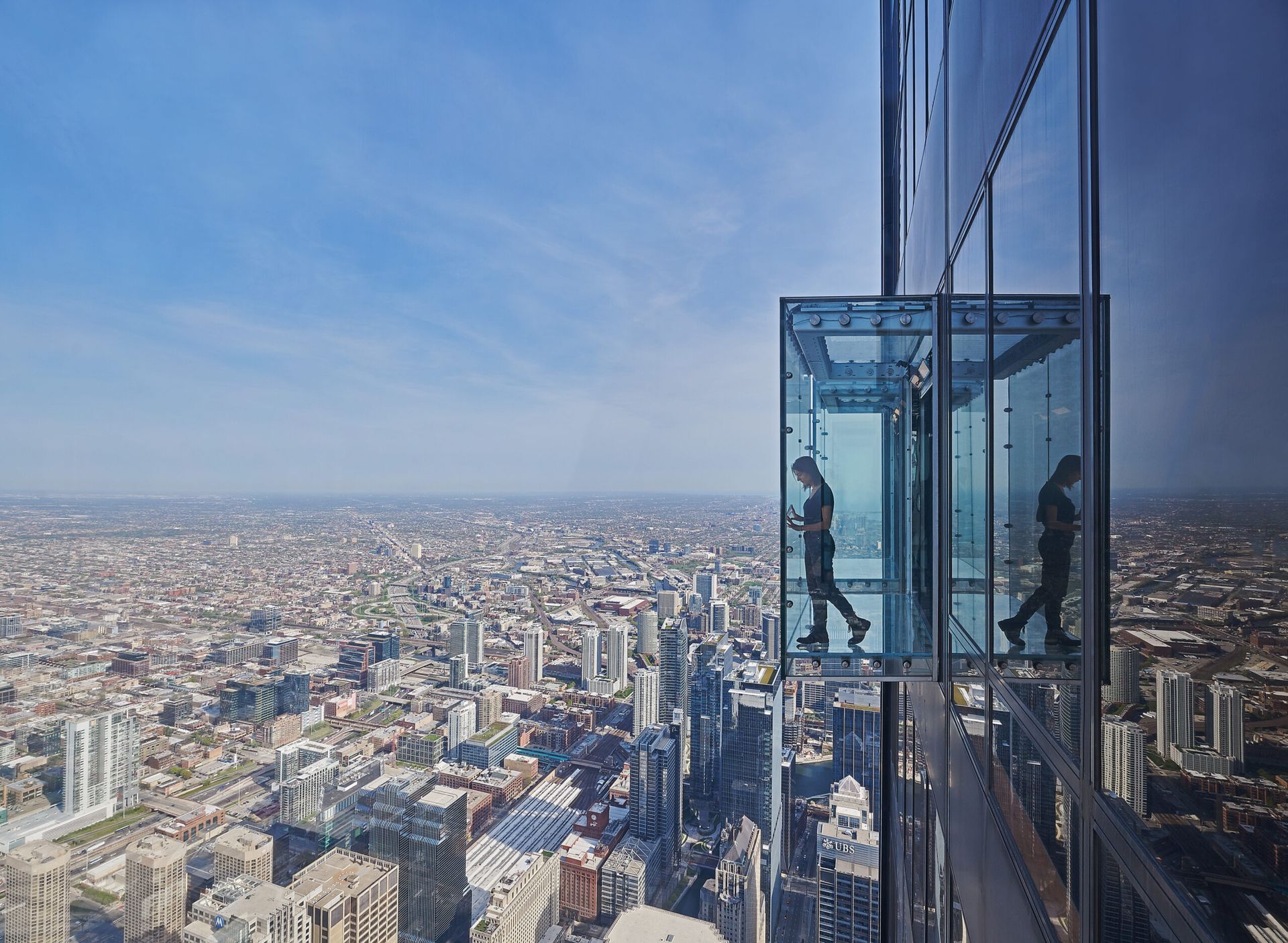 Two people are walking on a glass platform on top of a tall building overlooking a city.