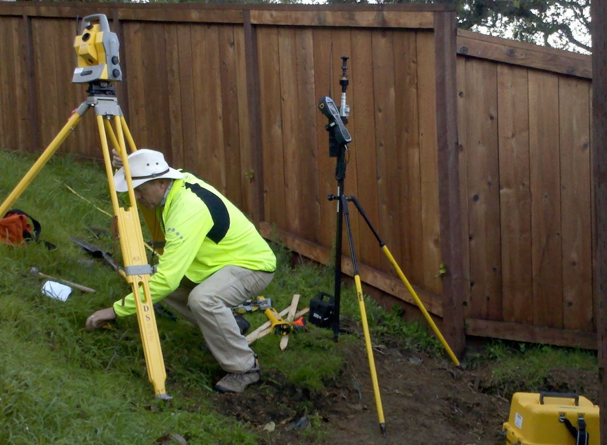 Person in safety vest using a surveying instrument on a tripod; outdoors near a wooden fence.