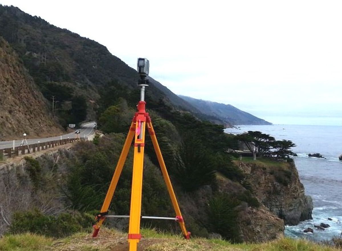 Surveying equipment on a tripod, set against a mountainous coastal backdrop with a road.
