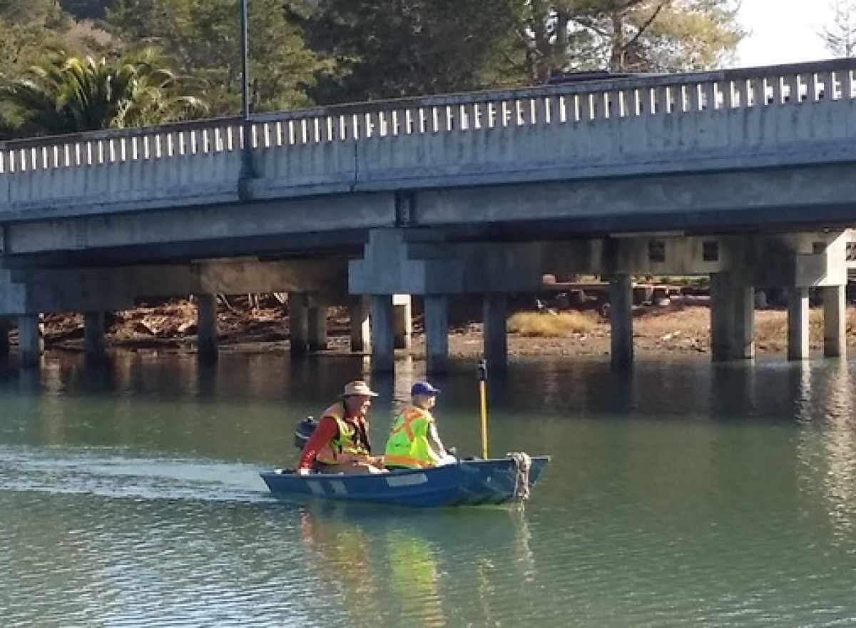 Two people in a blue boat under a bridge, one wearing a vest, on the water.