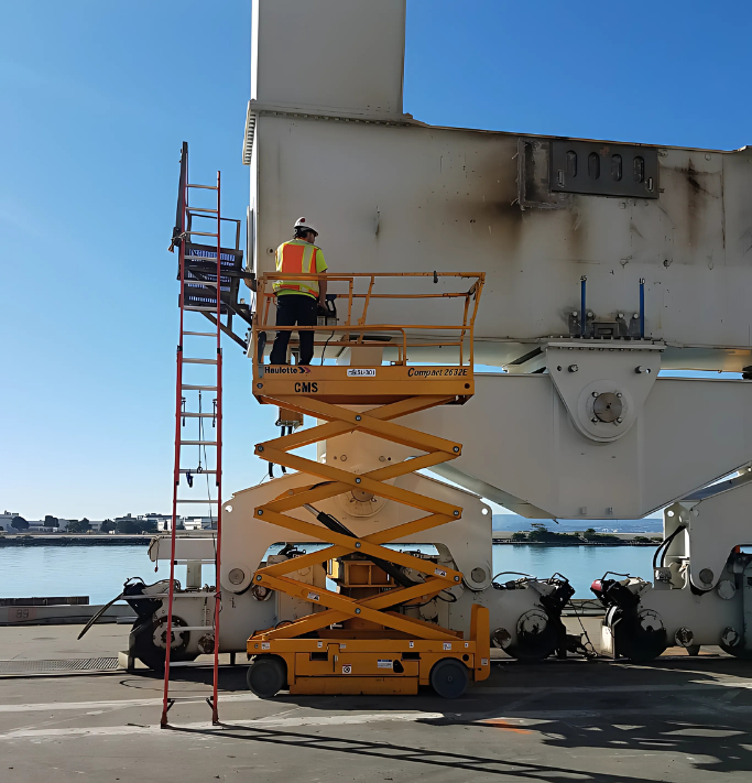 Worker on yellow lift platform near large white structure, red ladder alongside. Outdoor setting, blue sky.