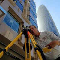 Man using surveying equipment, measuring a building with a tall skyscraper in the background. Sunny day.