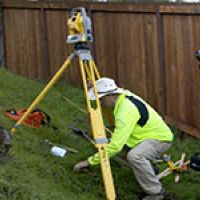 Person in safety vest using a surveying instrument on a tripod; outdoors near a wooden fence.