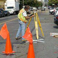 Surveyor in a street using a tripod-mounted instrument, near orange cones and flags, measuring for road work.