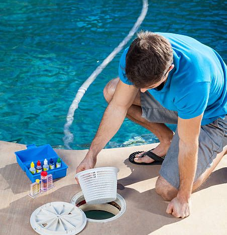 Man kneeling by pool skimmer, placing basket into open drain beside pool cleaning supplies.