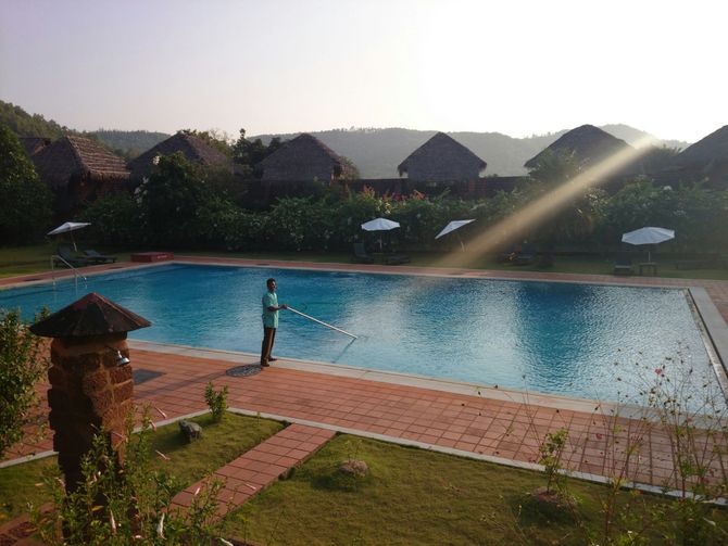 Resort pool at sunrise with a person standing on the deck and lounge umbrellas beyond the water