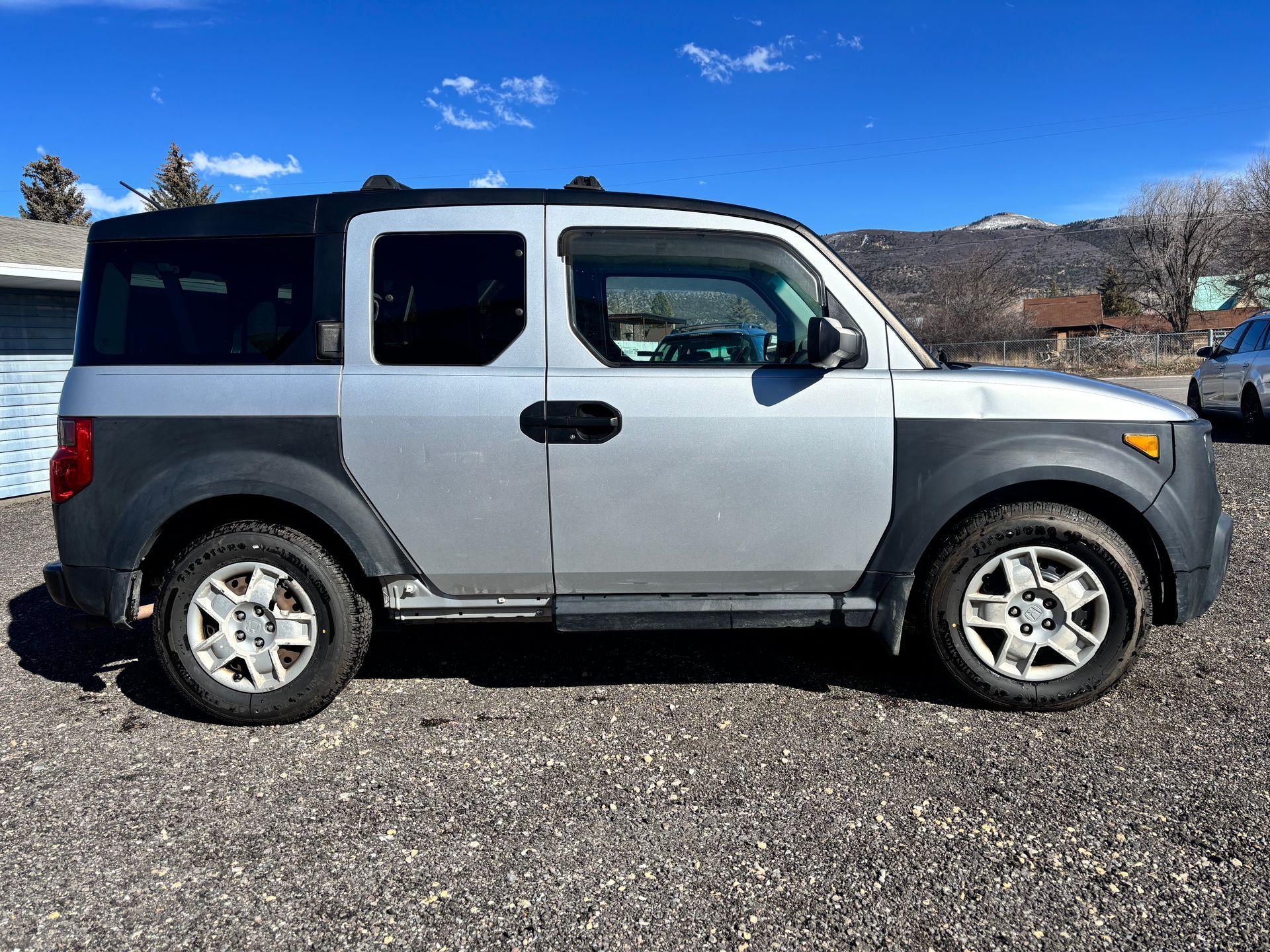 Gray two-door SUV parked on gravel under a blue sky with mountains in the background