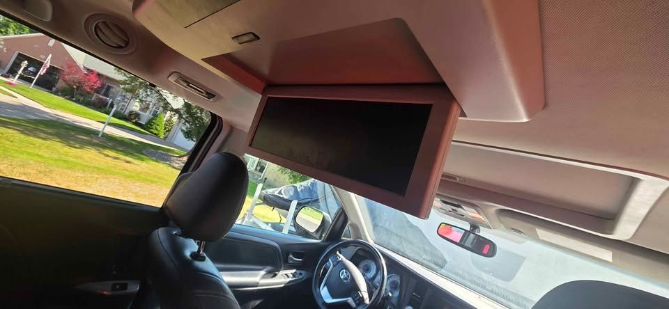 Car interior with a large overhead screen, front seat, and view through the windshield to parked cars outside.