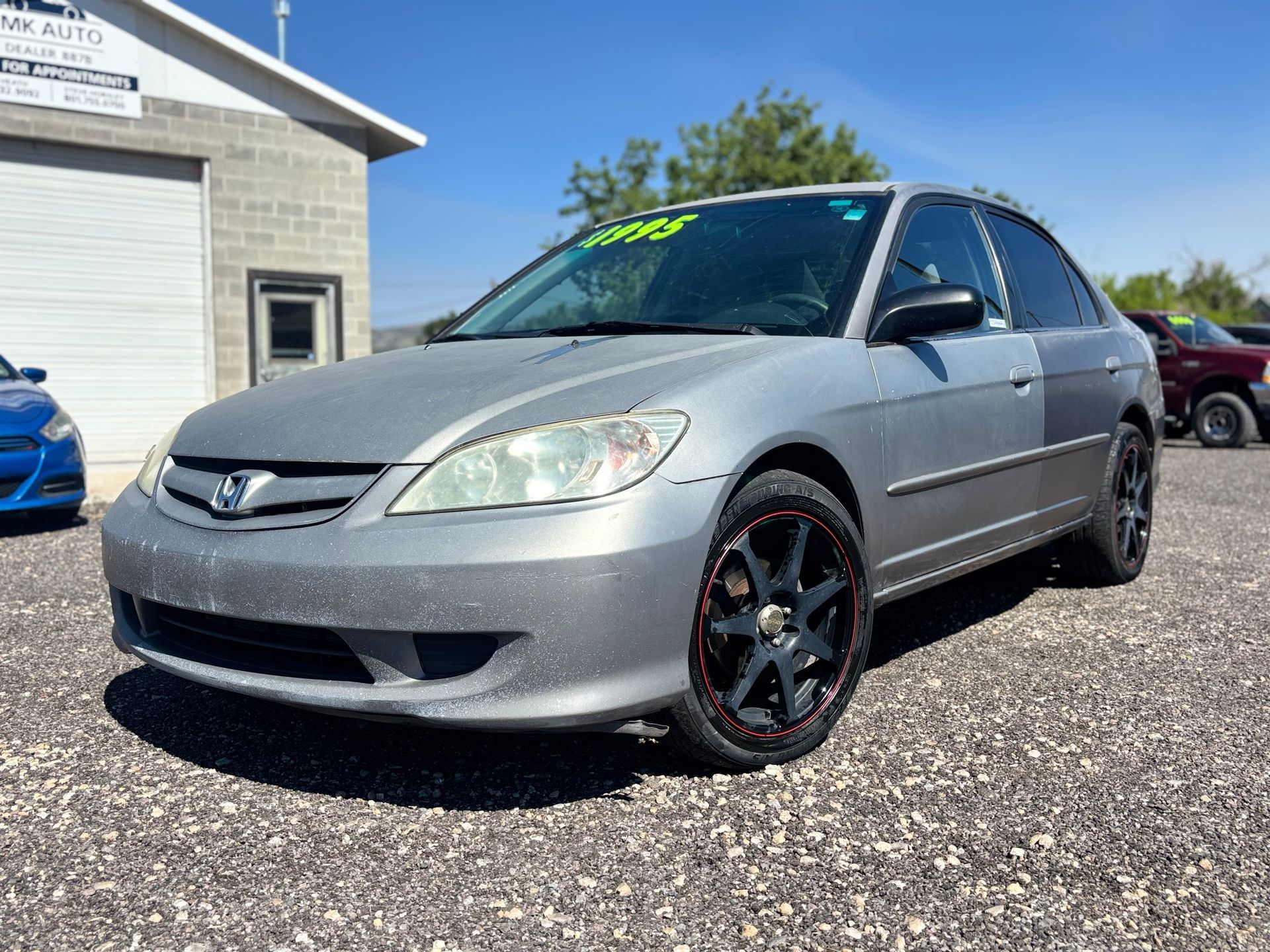Silver Honda sedan parked outside a building on a gravel lot, with black and red rims.
