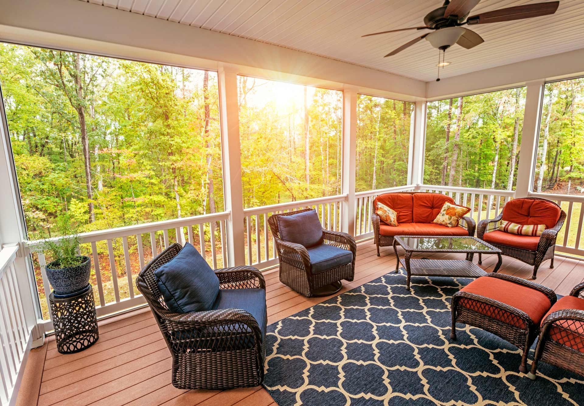 Screened porch with wicker furniture, blue rug, and view of trees and sunlight.