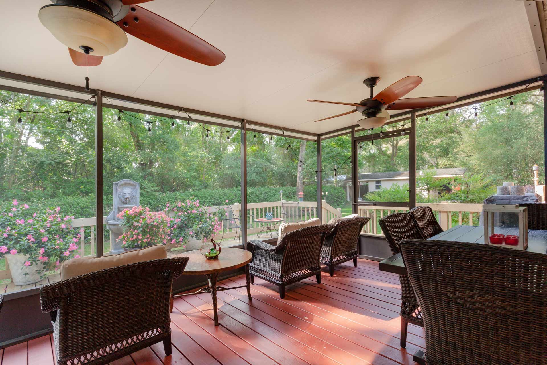 Screened porch with wicker furniture, string lights, and ceiling fans overlooking a wooden deck and garden.