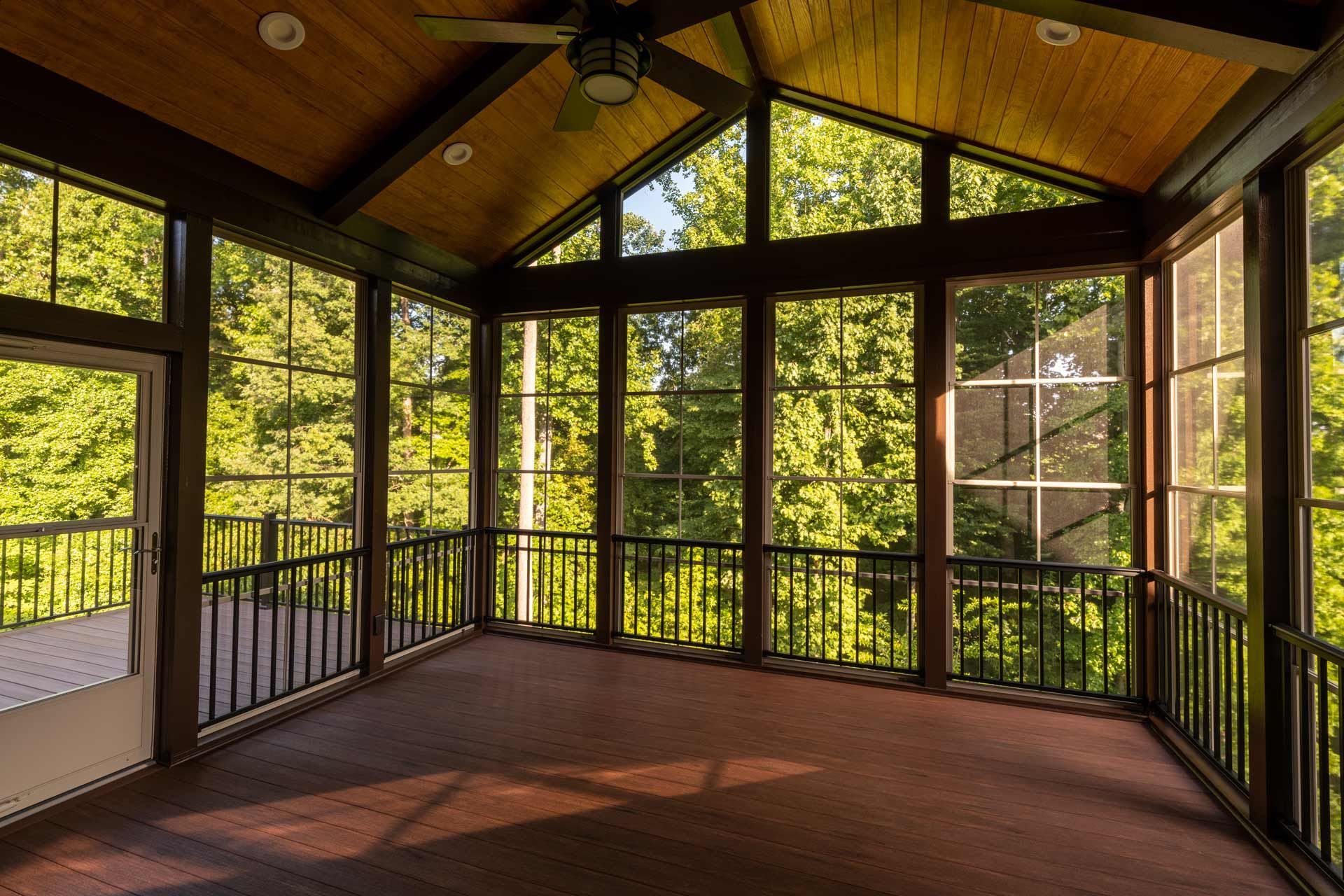 Screened-in porch with dark wood trim and railing, filled with sunlight and views of lush trees.